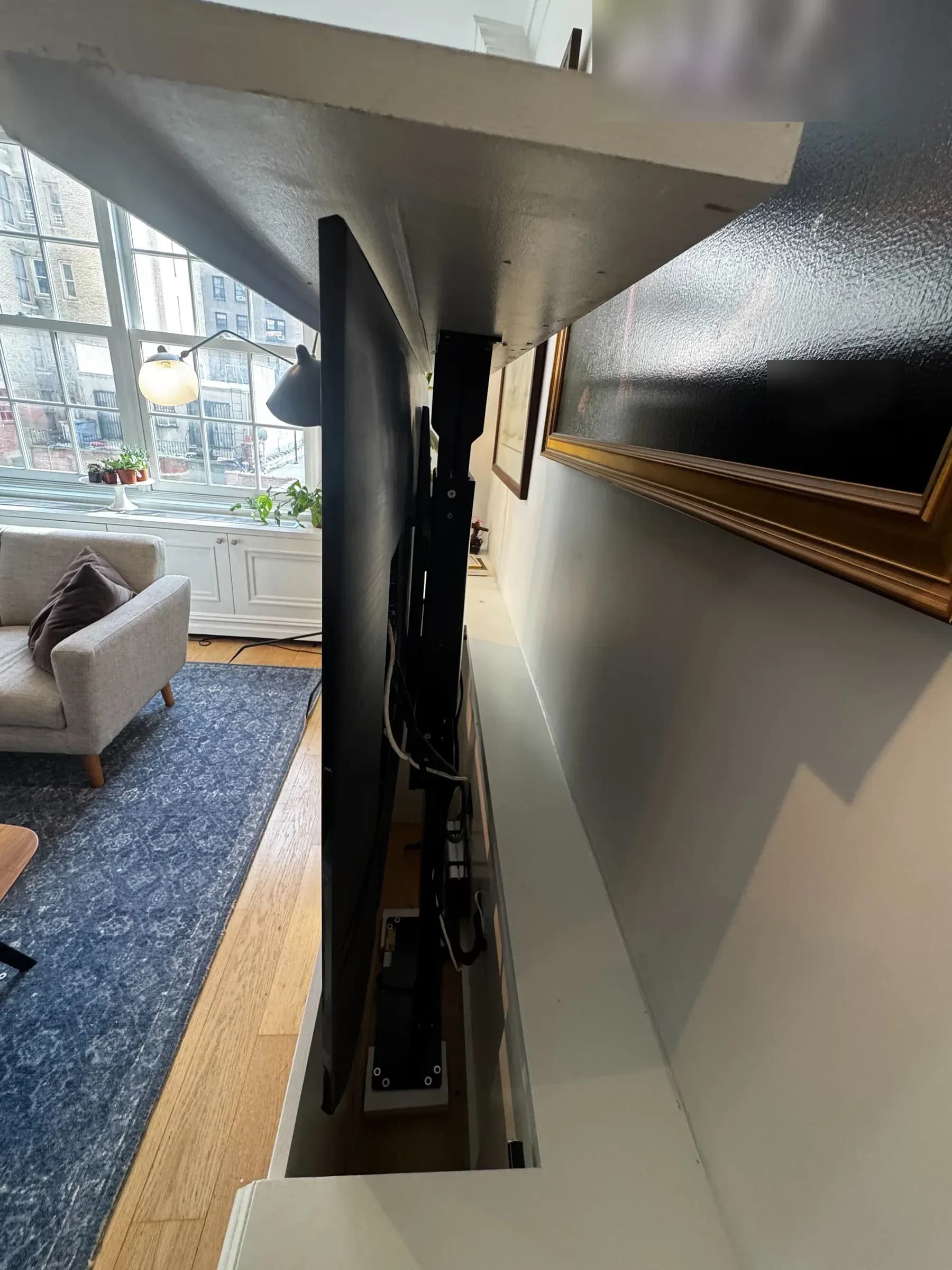 A side view of the motorized TV lift mechanism behind the TV, with a window and a framed picture visible nearby.