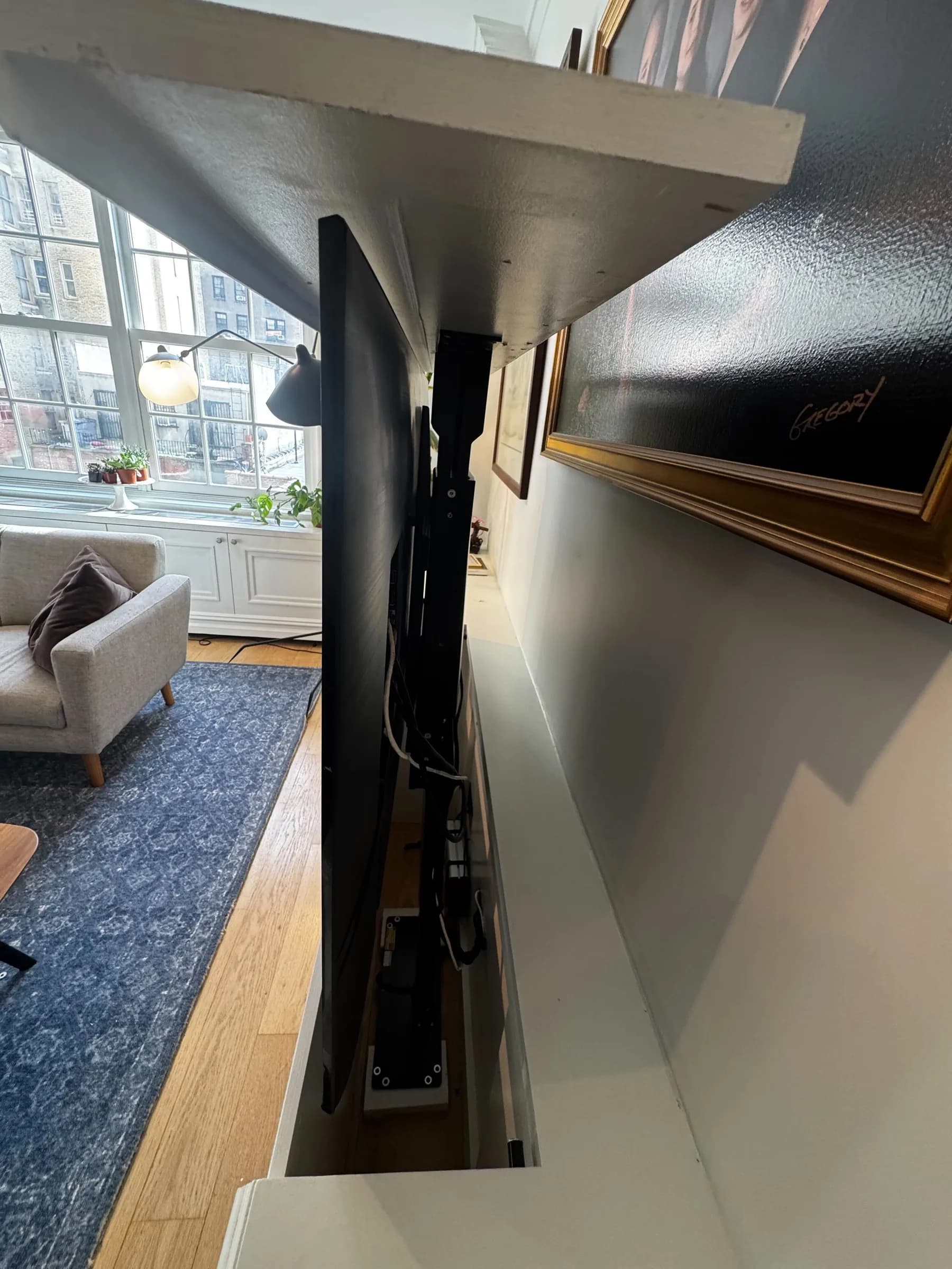A side view of the motorized TV lift mechanism behind the TV, with a window and a framed picture visible nearby.