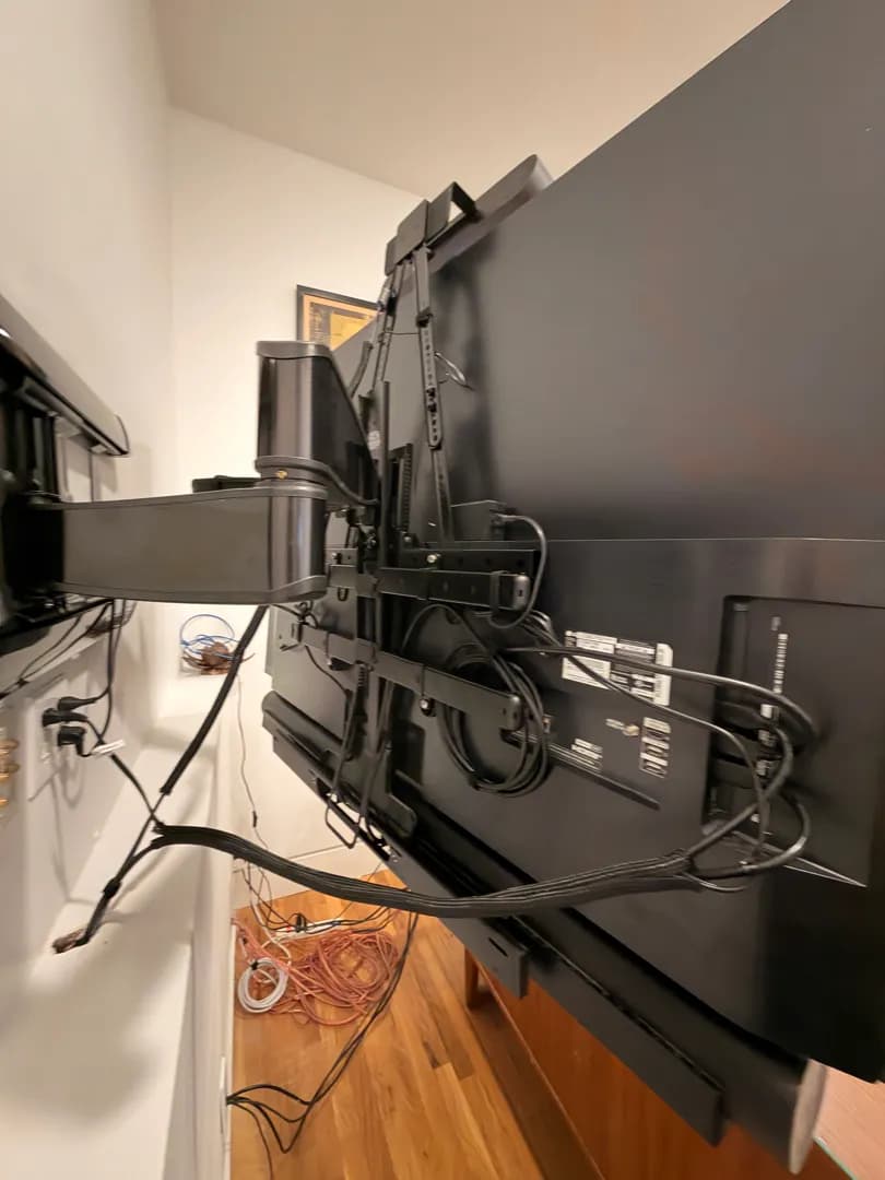 A close view from behind the TV showing the Sanus full-motion mount extended from the wall, with the VESA brackets and cables.
