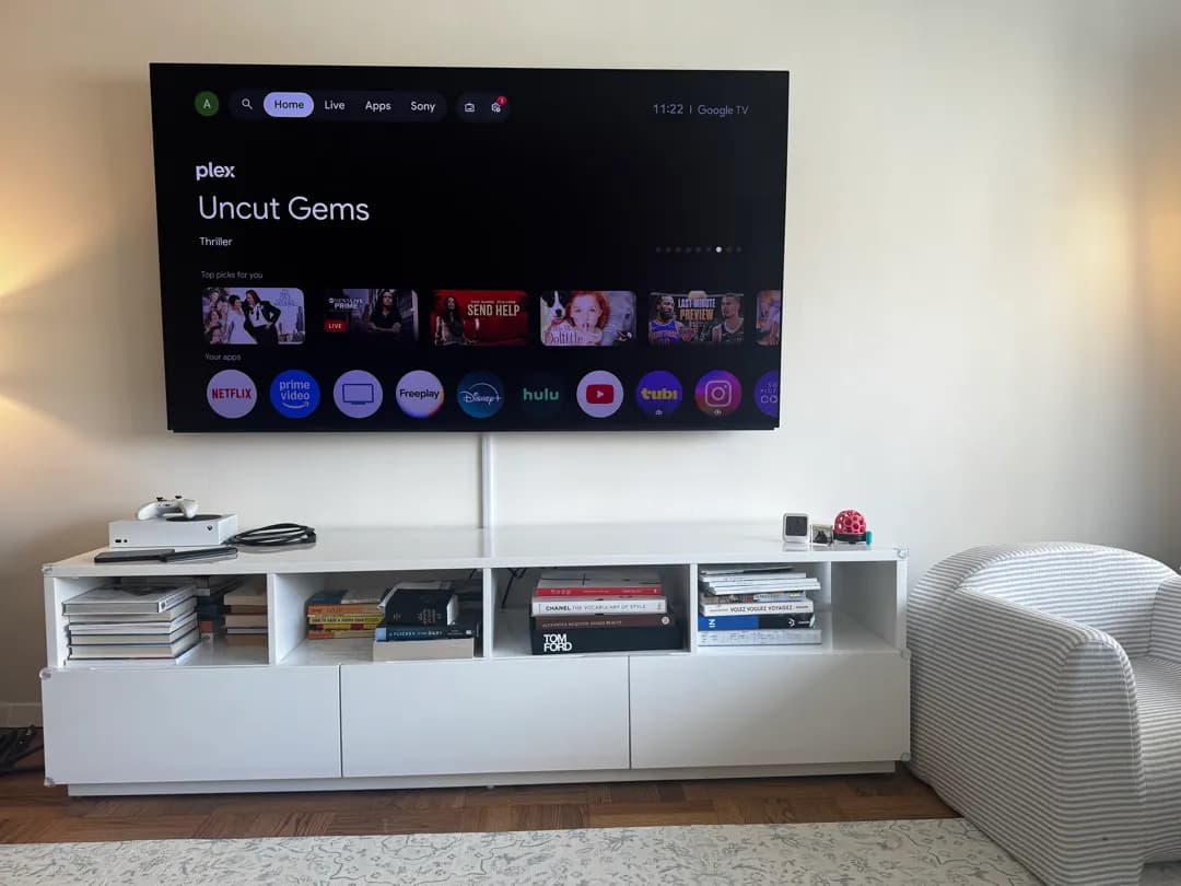 A wall-mounted TV shows a streaming home screen above a white media console with books and a striped armchair nearby.